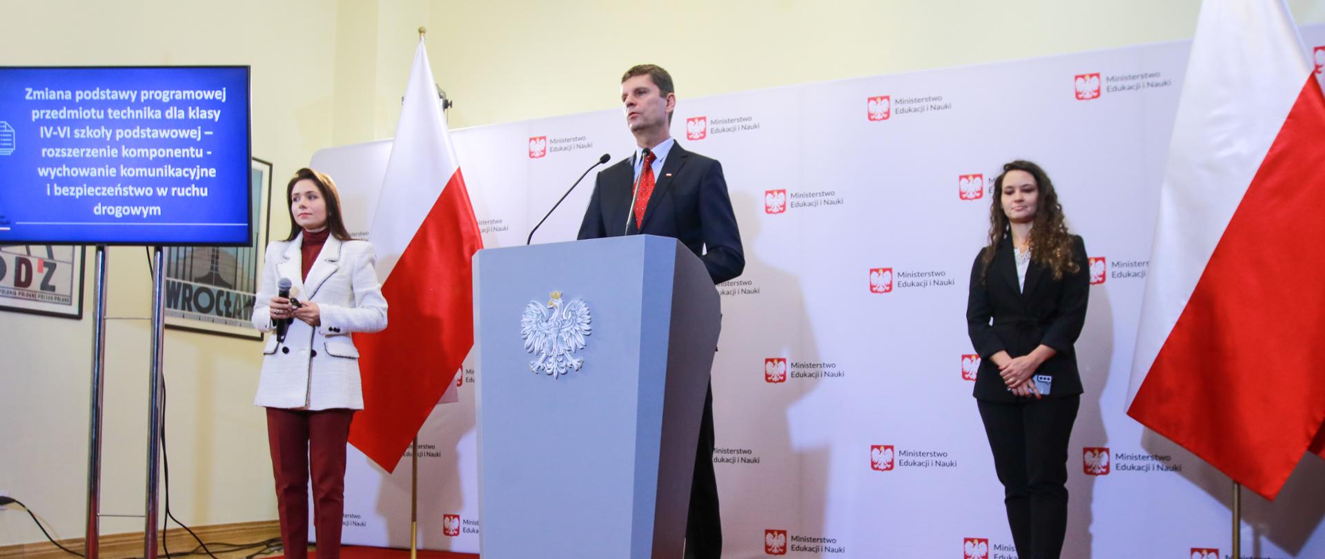Wiceminister Dariusz Piontkowski stoi przy mównicy, po lewej stoi rzecznik prasowy, po porawej Justyna Orłowska. Czerwony dywan, w tle ścianka z logotypem Ministerstwa Edukacji i Nauki 