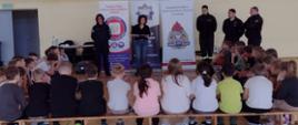 Zdjęcie przedstawia zajęcia edukacyjne w sali gimnastycznej, gdzie dwóch dorosłych, w tym jedna kobieta i jeden mężczyzna, objaśnia coś grupie dzieci siedzących na ławkach. W tle widoczne są dwa banery, jeden należący do Powiatowej Stacji Sanitarno-Epidemiologicznej w Zwoleniu, a drugi do Komendy Powiatowej Państwowej Straży Pożarnej w Zwoleniu. Na prawym banerze widnieje logo PSP. Obok banerów stoją trzej mężczyźni w mundurach strażackich. Sala jest jasna, z drewnianą podłogą.