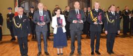 Dzień Strażaka w Rawiczu. Sala widowiskowa w domu kultury. Na tle pododdziału w szeregu stoją: z-ca komendanta powiatowego PSP, ministrowie, którzy otrzymali okolicznościowe statuetki w podziękowaniu za wsparcie komendy, komendant wojewódzki PSP i komendant powiatowy PSP.