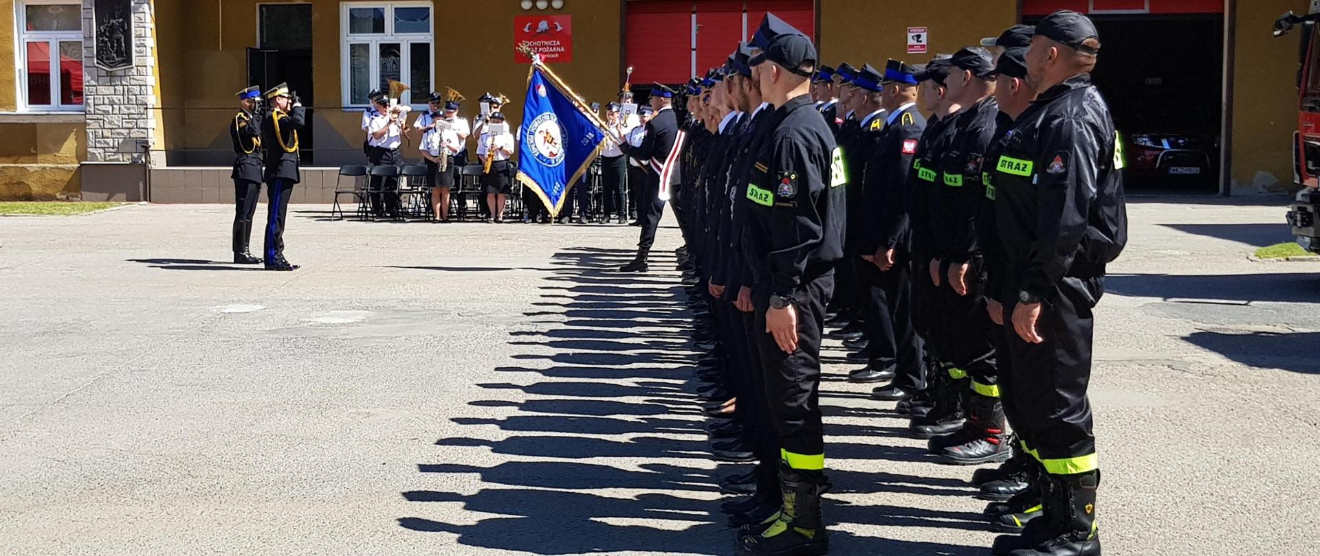strażacy na placu stoją w dwuszeregu, w tle orkiestra dęta, przed strażakami komendant z dowódcą uroczystości salutują sztandarowi