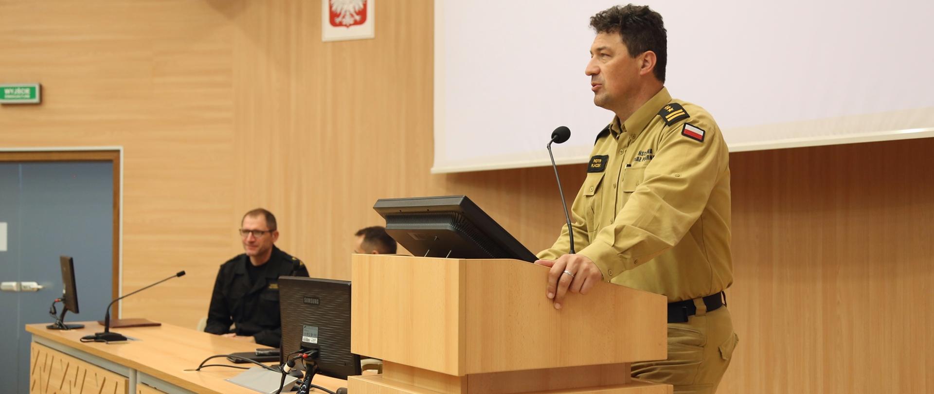 Komendant Centralnej Szkoły PSP przemawia stojąc na podeście przy mównicy, w głębi przy stole siedzący strażak
