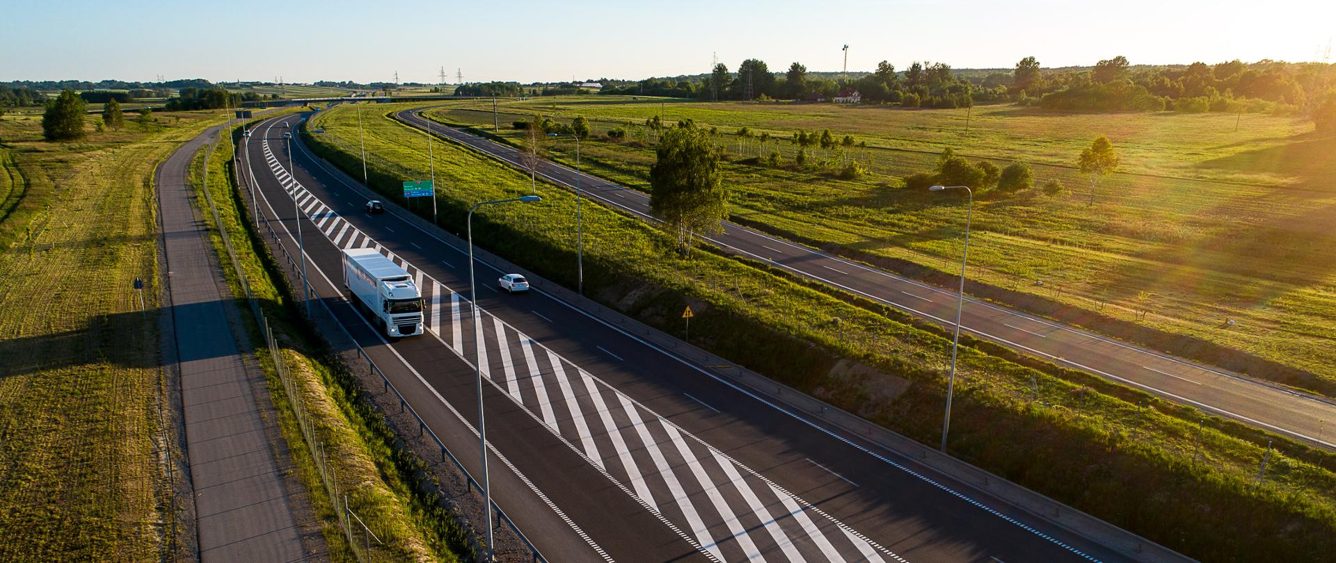 Widok na drogę ekspresową z lotu ptaka. Na jezdni jadą auta w tym ciężarowe. Dookoła zieleń.