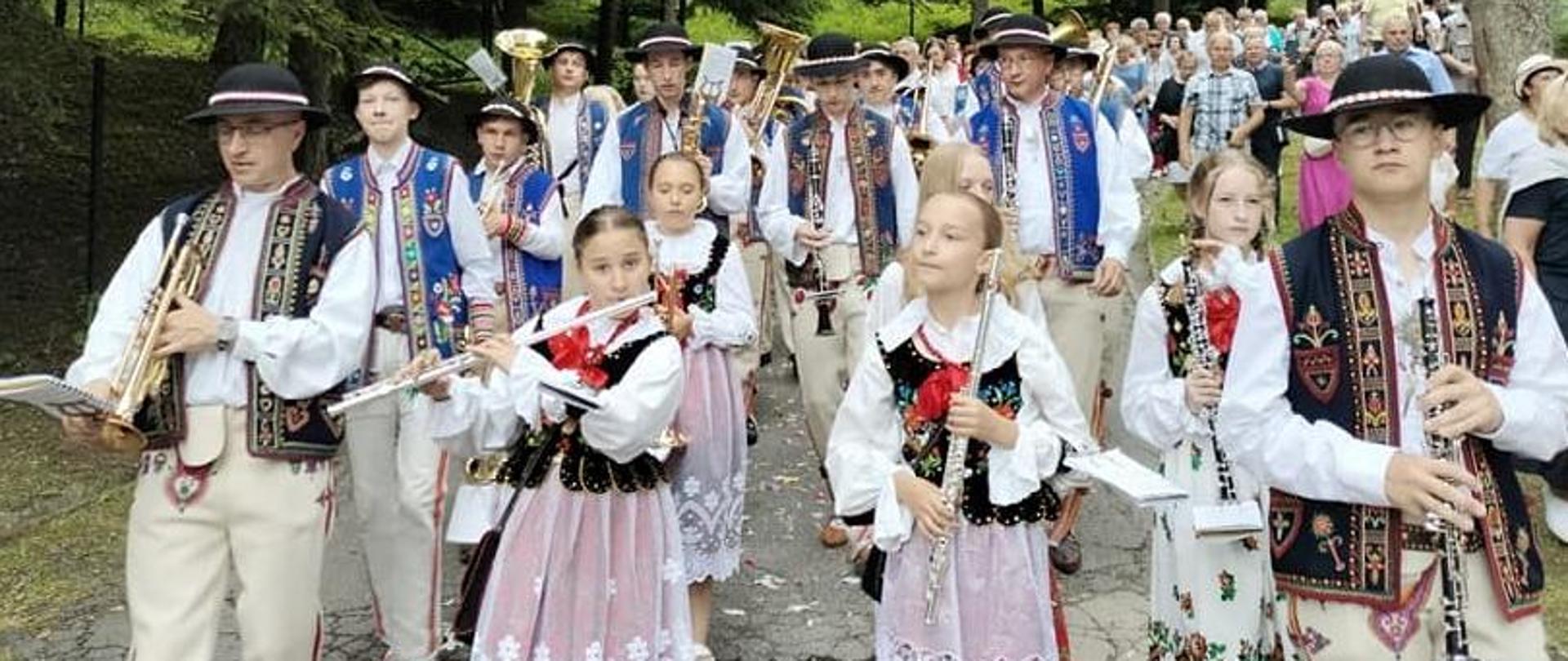 Procesja Bożego Ciała w terenie leśnym, uczniowie ubrani po góralsku, grający na instrumentach idą wraz z procesją Bożego Ciała.