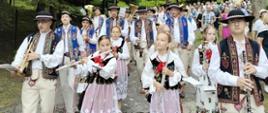 Procesja Bożego Ciała w terenie leśnym, uczniowie ubrani po góralsku, grający na instrumentach idą wraz z procesją Bożego Ciała.