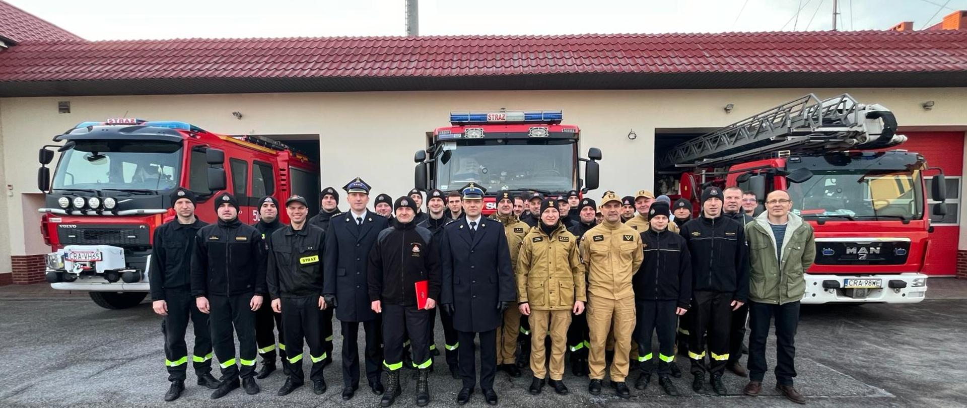 Zdjęcie przedstawia strażaków PSP podczas uroczystej zmiany służby w związku z przejściem na zaopatrzenie emerytalne Marcina. Na drugim planie pojazdy pożarnicze.