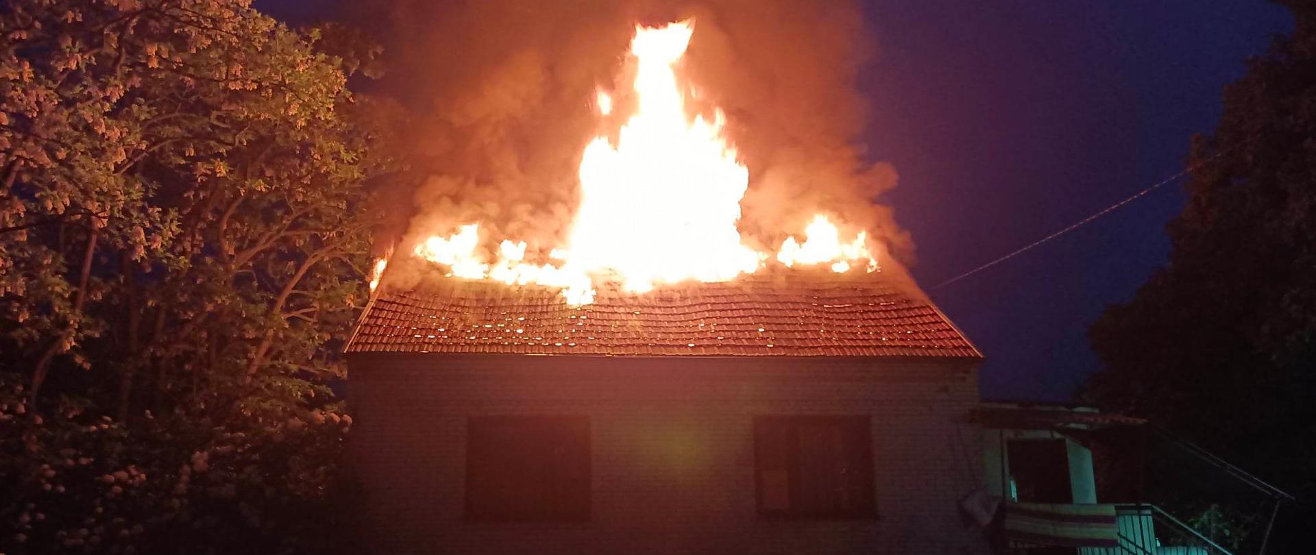 Dom parterowy jednorodzinny na którym widać wydobywające się przez dach płomienie. Na zdjęciu jest ciemno, po lewej stronie widać drzewo.