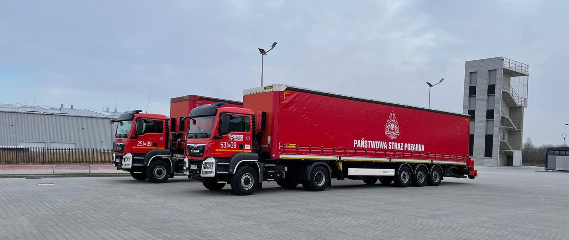 Zdjęcie przedstawia dwa tiry transportowe Państwowej Straży Pożarnej ustawione na placu wewnętrznym Jednostki Ratowniczo-Gaśniczej nr 3 w Rzeszowie