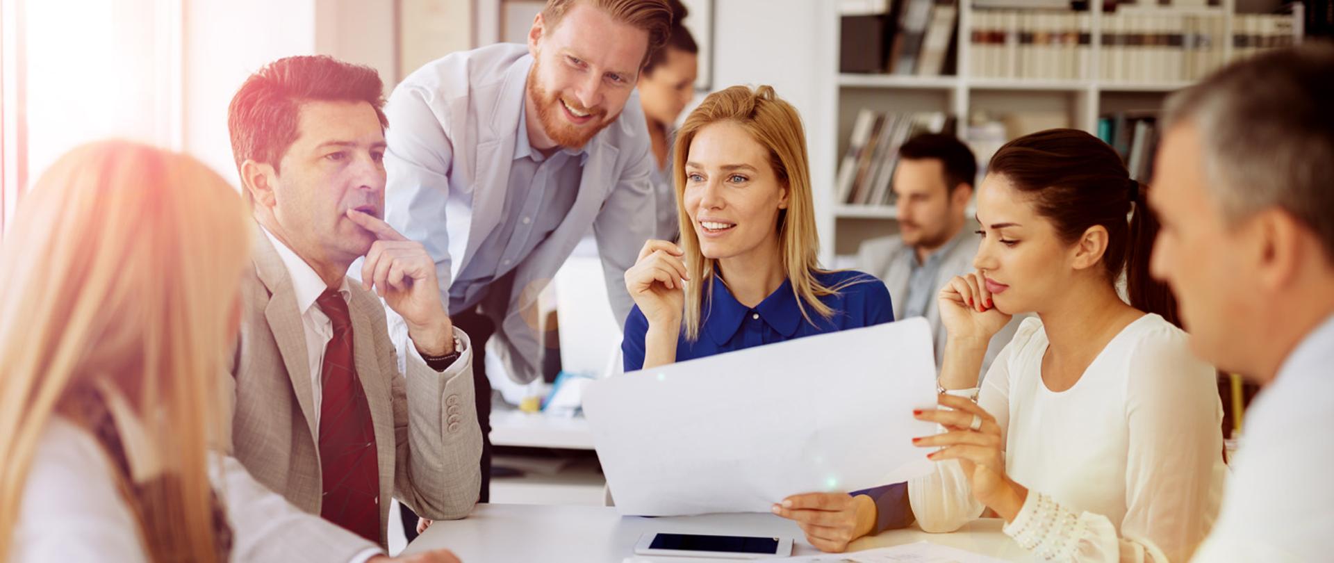 grupa pracowników siedzących wokół stołu rozmawia o propozycji na kartce trzymanej przez jedną z osób