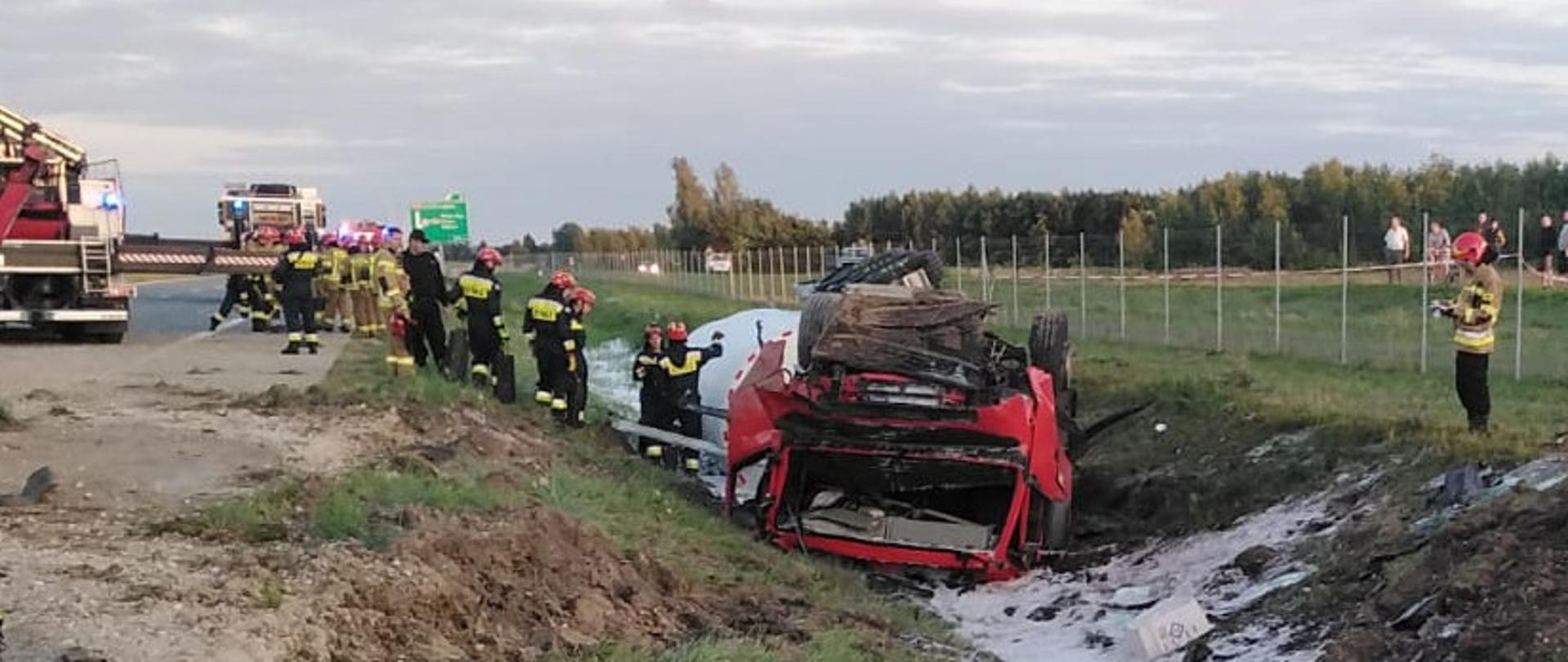 Na zdjęciu uszkodzony ciągnik siodłowy w rowie. Za nim uszkodzona cysterna również w rowie. Na lewo od ciągnika siodłowego strażacy 
