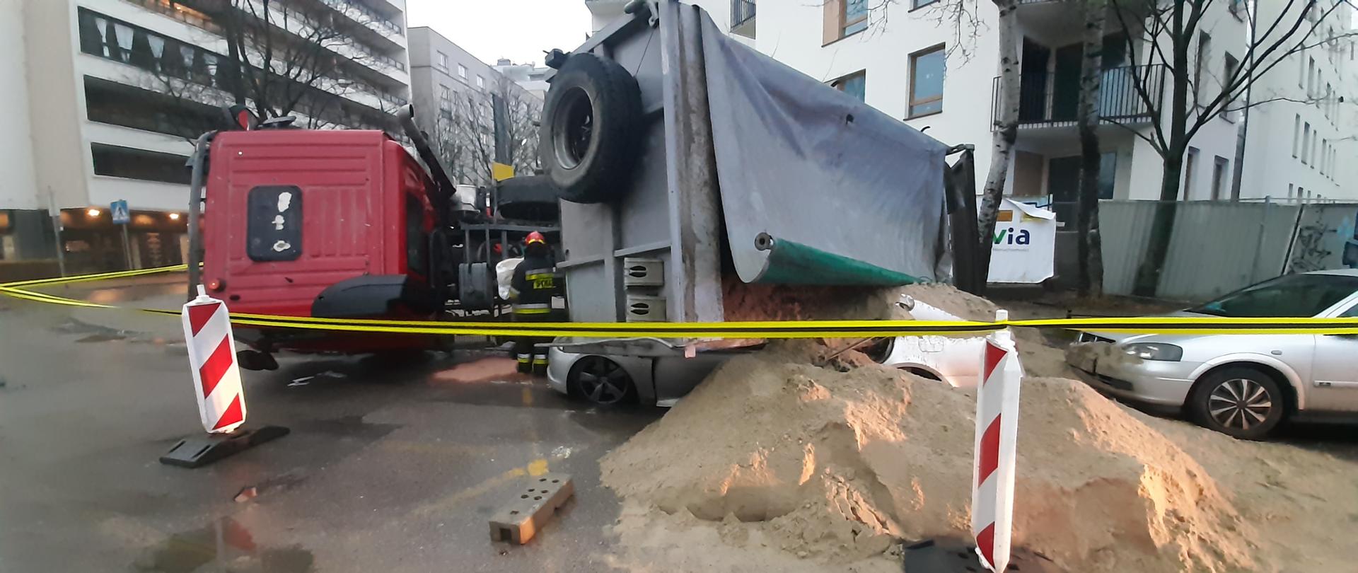 Na zdjęciu widać przewrócony samochód ciężarowy , który przewrócił się ma samochód osobowy. Piach wysypał się z samochodu ciężarowego i leży na ulicy. Miejsce zdarzenia zabezpieczone jest czerwono - białymi pachołkami oraz żółto- czarną taśmą ostrzegawczą.