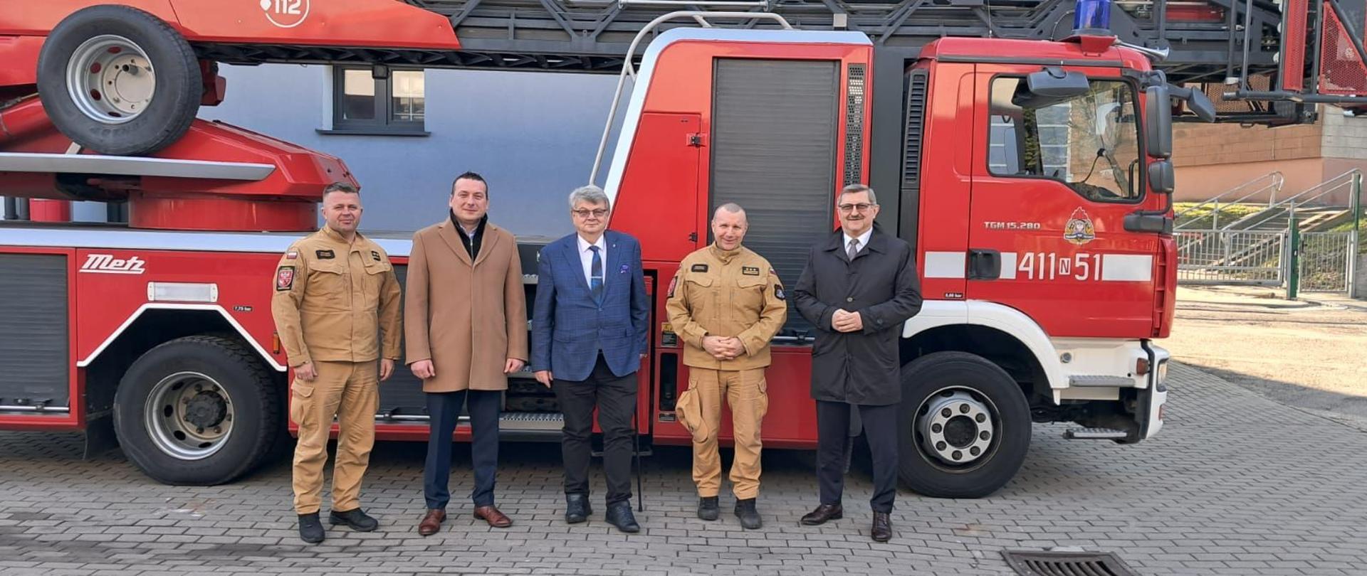 Na tle mechanicznej drabiny strażackiej stoi pięć osób. Dwóch funkcjonariuszy PSP oraz trzech członków zarządu powiatu braniewskiego.