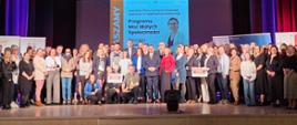 Zdjęcie grupowe uczestników konferencji. W tle ekran z informacją: Sekretarz Stanu Adriana Porowska zaprasza na regionalną konferencję Programu Moc Małych Społeczności.