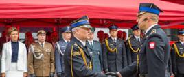 Zastępca Mazowieckiego Komendanta Wojewódzkiego PSP st. bryg. Waldemar Wysowski (w centrum, z profilu) przyjmuje meldunek o zakończeniu uroczystości z okazji Dnia Strażaka w Siedlcach. W tle, pod czerwonym namiotem ogrodowym, stoi grupa zaproszonych gości, w tym kobieta w białym żakiecie, żołnierz w oliwkowym mundurze, policjant oraz pozostali strażacy w umundurowaniu galowym.