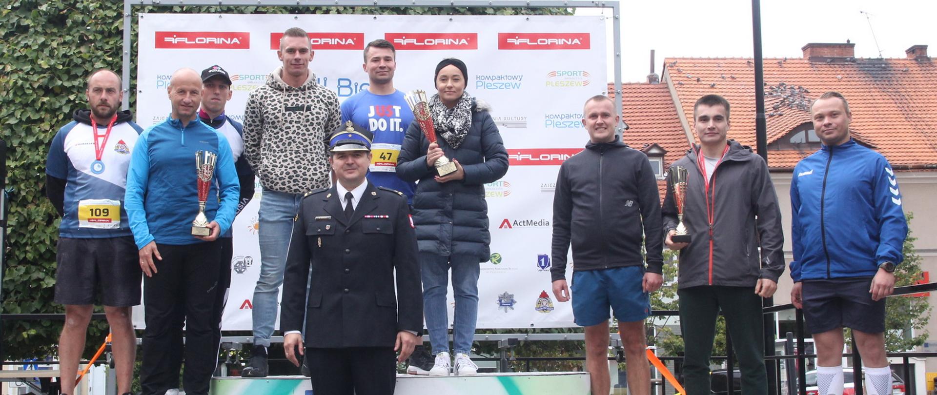 biegacze stoją na podium, trzymaja puchary, przed podium stoi strażak w rogatywce i mundurze wyjściowym