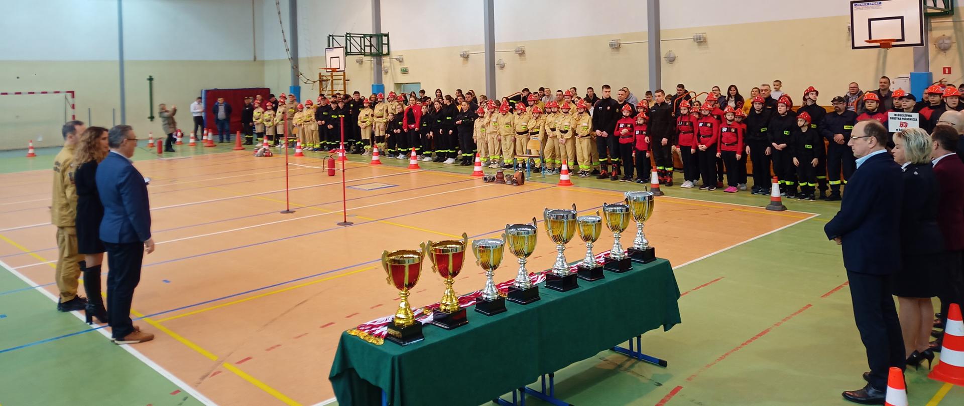 Zdjęcie przedstawia wnętrze hali sportowej w Sabniach. Jest to moment zbiórki zawodników MDP, przedstawicieli władz samorządowych oraz przedstawiciela KP PSP Sokołów Podlaski. Na pierwszym palnie nagrody za poszczególne miejsca. W dalszej części zawodnicy w grupach poszczególnych drużyn z terenu powiatu.