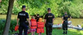 Nad brzegiem rzeki strażacy, policjanci oraz dwójka dzieci przy łodzi ratowniczej