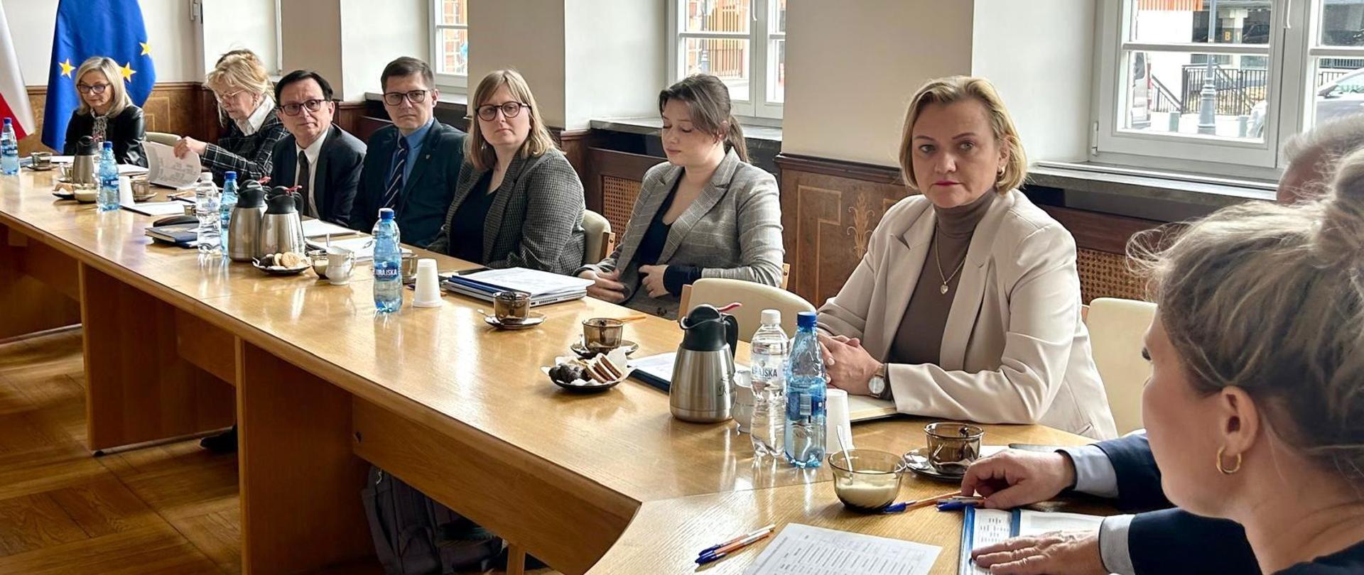 Kilka osób siedzi przy okrągłym stole i słucha przemawiającej kobiety. 