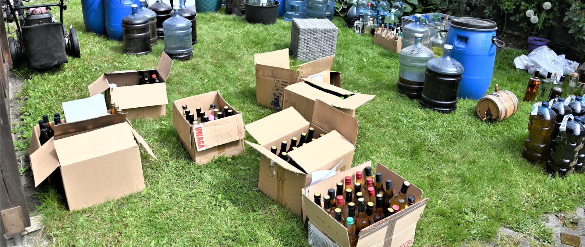 Butelki i beczki z bimbrem stoją na trawie.
