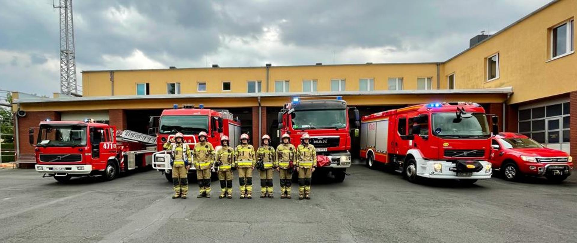 Na pierwszym planie strażacy zmiany służbowej, w tle samochody pożarnicze oraz garaż.