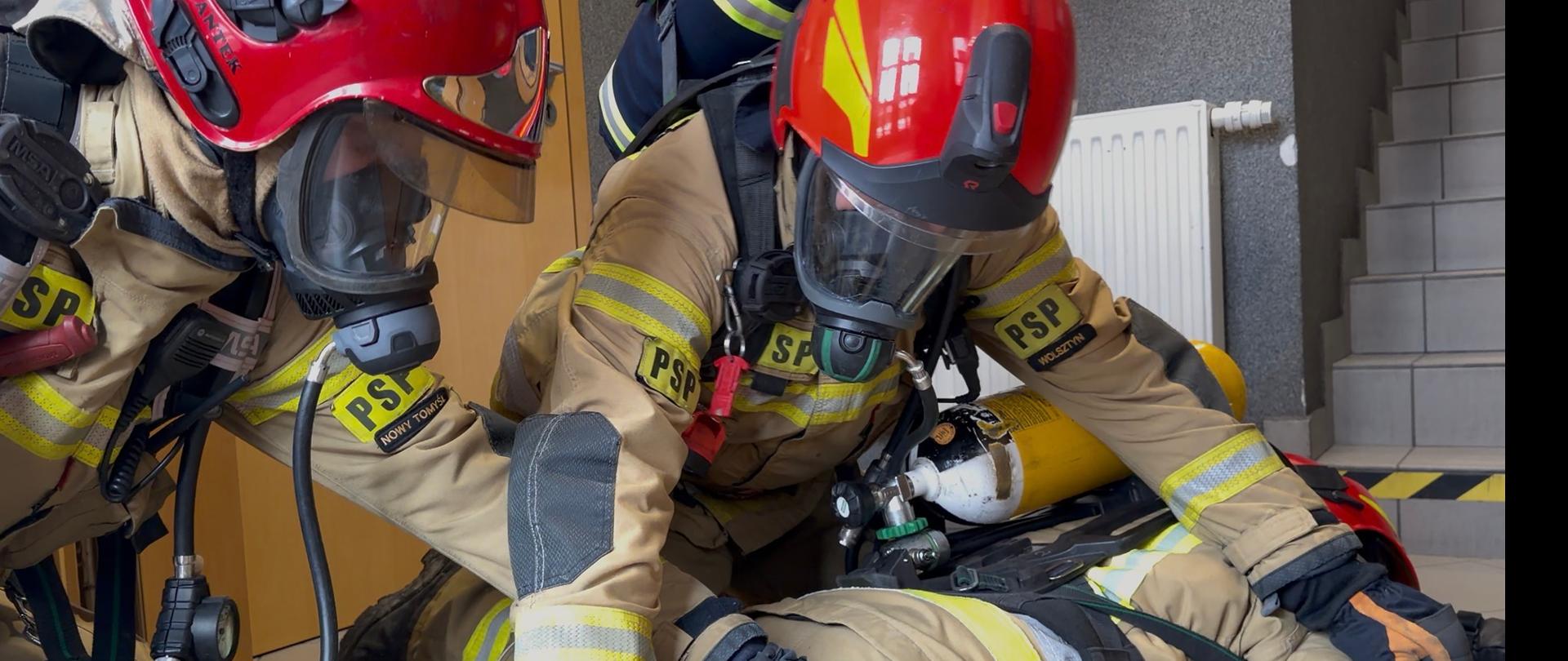 Na zdjęciu dwóch strażaków przekłada trzeciego leżącego na ziemi strażaka w pozycję bezpieczną do ewakuacji. 