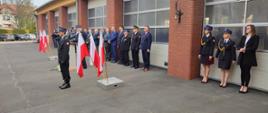 Na placu strażackim przy bramach garażowych koloru białego stoją strażacy i osoby cywilne. Przed osobami znajdują się w stojakach flagi biało-czerwone