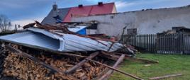 Skutki orkanu Dudley. Dąbrówka - teren jednej z posesji. Na stercie drewna leży zerwany z pobliskiego budynku gospodarczego. Pokrycie z blachy jest zwinięte. W głębi budynek.