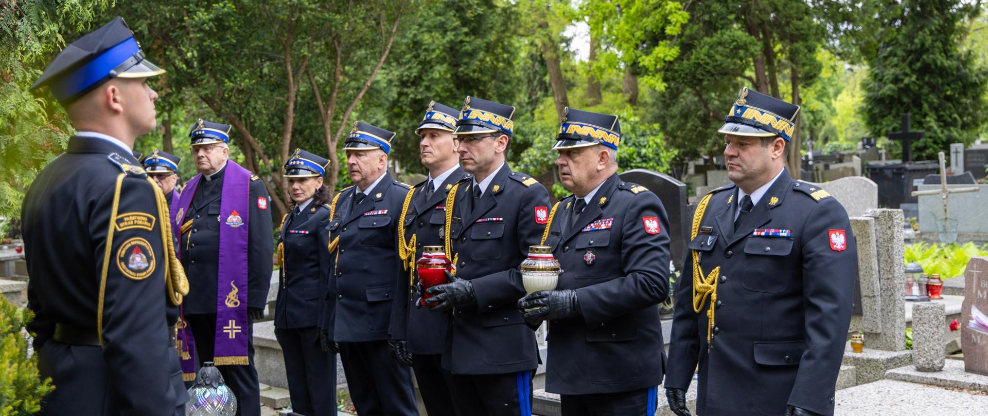 Komendant główny PSP wraz z kapelanami oraz innymi dowódcami straży pożarnej w szeregu podczas uroczystości przy grobach zasłużonych strażaków. Wszyscy ubrani w mundury galowe, część w stułach liturgicznych.