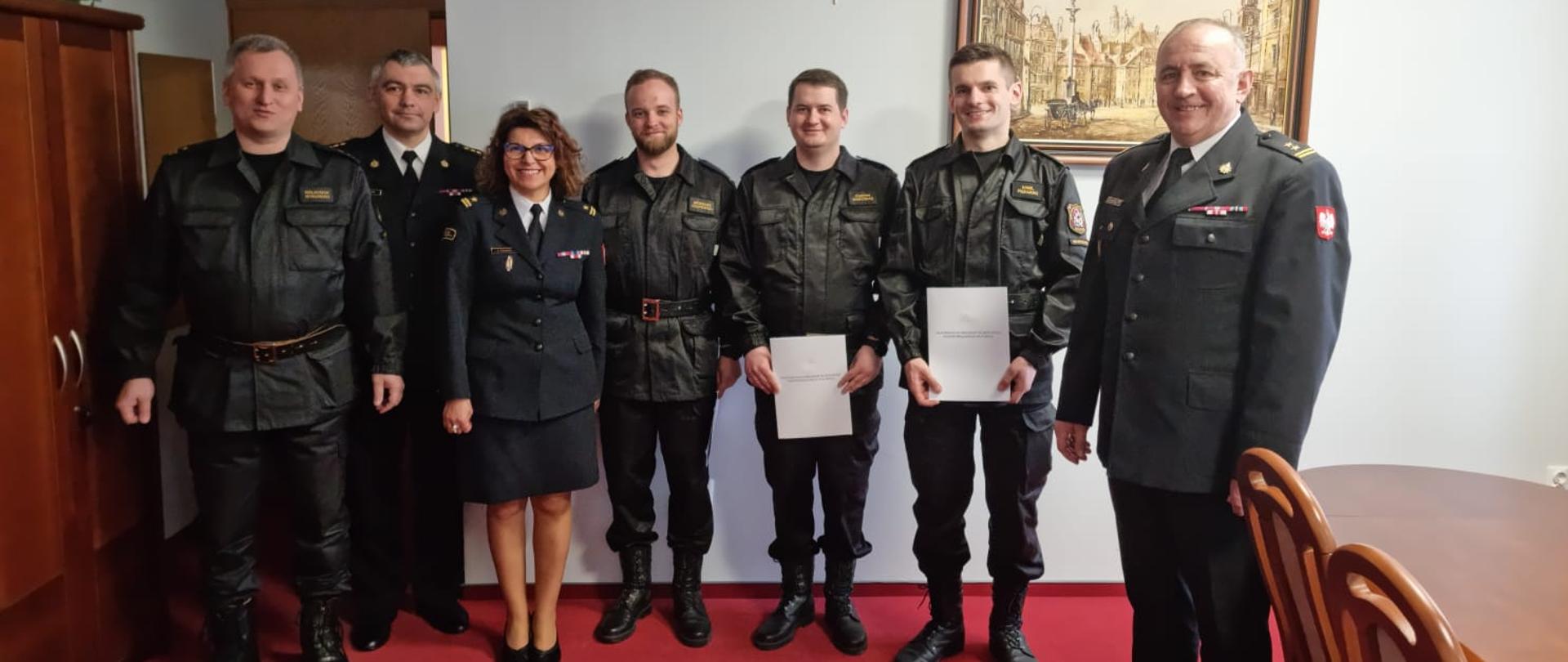 Na zdjęciu w gabinecie Komendanta Miejskiego PSP m.st. Warszawy stoją od lewej: młodszy brygadier Waldemar Wysowski. starszy brygadier Artur Rudzik, młodszy brygadier Anna Zielińska, starszy strażak Grzegorz Czapiewski, starszy strażak Damian Marciniak, starszy strażak Kamil Piekarski, brygadier Roman Krzywiec. Starsi strażacy trzymają w rękach teczki z wręczonymi dokumentami. 