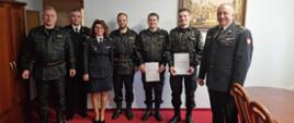 Na zdjęciu w gabinecie Komendanta Miejskiego PSP m.st. Warszawy stoją od lewej: młodszy brygadier Waldemar Wysowski. starszy brygadier Artur Rudzik, młodszy brygadier Anna Zielińska, starszy strażak Grzegorz Czapiewski, starszy strażak Damian Marciniak, starszy strażak Kamil Piekarski, brygadier Roman Krzywiec. Starsi strażacy trzymają w rękach teczki z wręczonymi dokumentami. 