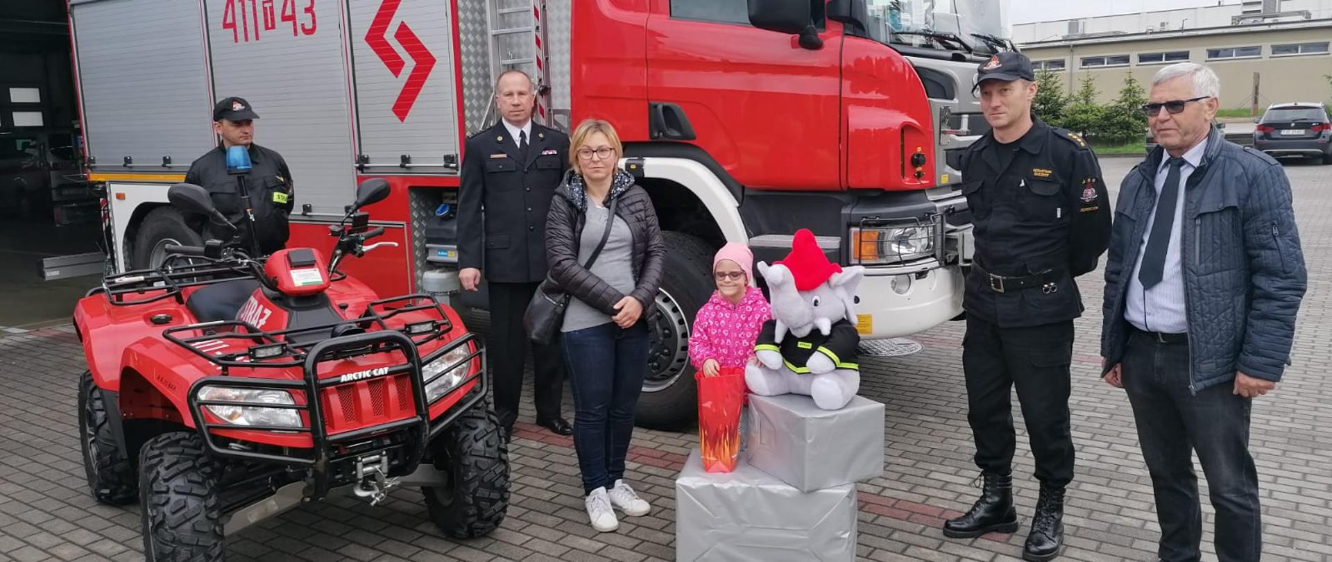 Zdjęcie przedstawia samochód specjalny ratownictwa technicznego marki Scania oraz quad'a. Na tle pojazdu ustawieni są; Komendant Powiatowy PSP w Jędrzejowie, dziewczynka wraz z opiekunami, dowódca Jednostki Ratowniczo-Gaśniczej w Jędrzejowie oraz pracownik Polskiego Związku Niewidomych. Przed dziewczynką ustawione są upominki, w tym pluszowy miś strażacki.
