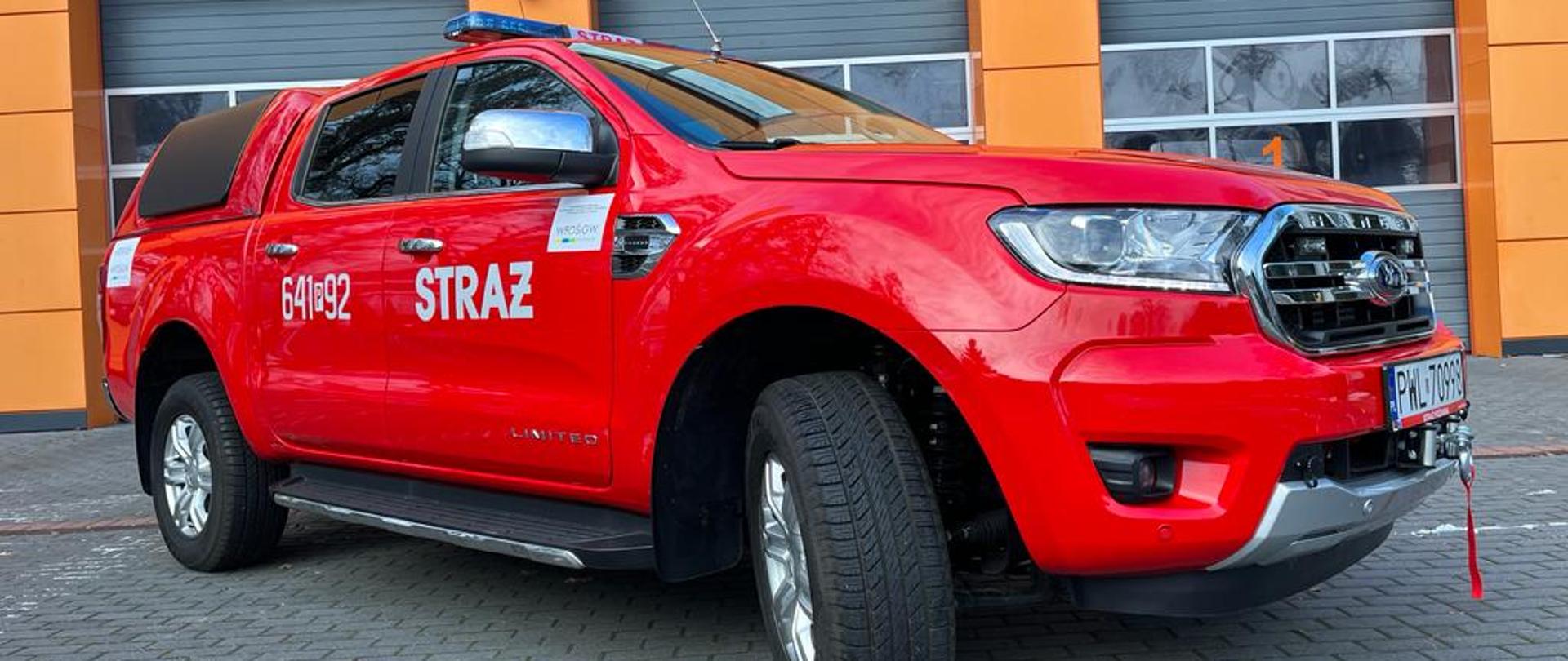 Zdjęcie przedstawia nowy czerwony samochód rozpoznawczo-ratowniczy marki Ford Ranger ustawiony bokiem na tle bram garażowych KP PSP w Wolsztynie. 