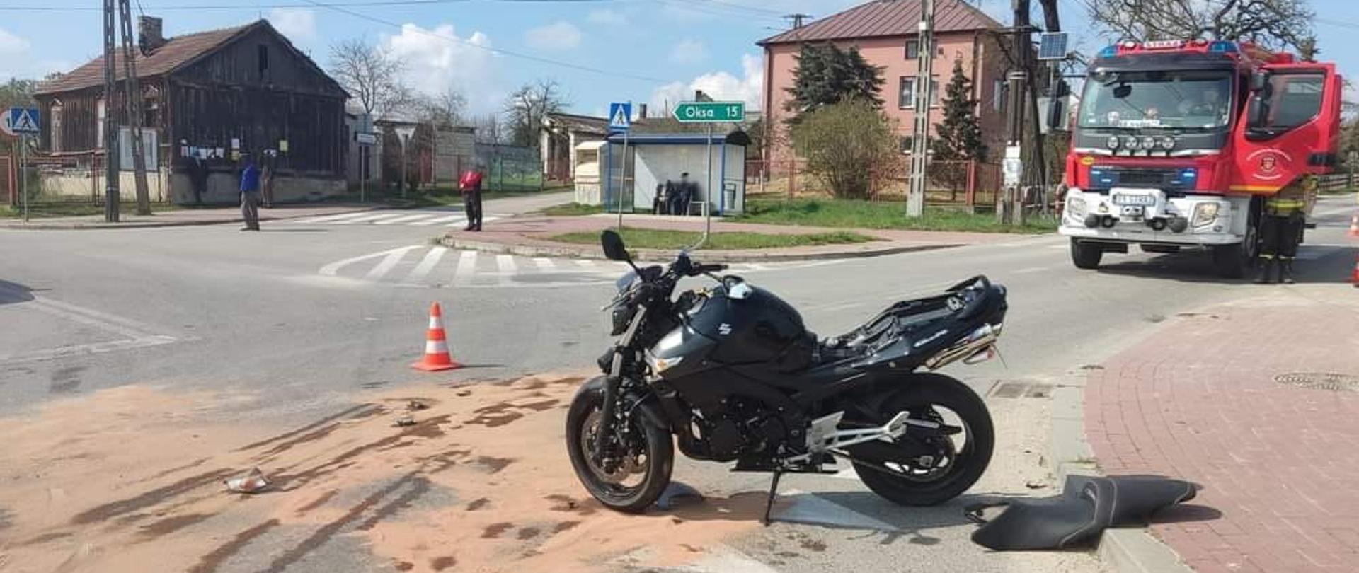 Zdjęcie przedstawia na pierwszym planie uszkodzony motocykl znajdujący się na skrzyżowaniu jezdni. Na drugim planie znajduje się samochód ratowniczo-gaśniczy. Wokół motocykla rozsypany sorbent neutralizujący wyciek płynów eksploatacyjnych.