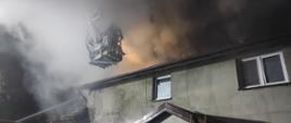 Zdjęcie przedstawia pożar domu mieszkalnego jednorodzinnego. Nad dachem unosi się silne zadymienie. Strażacy z kosza drabiny podają wodę na palący się budynek. 