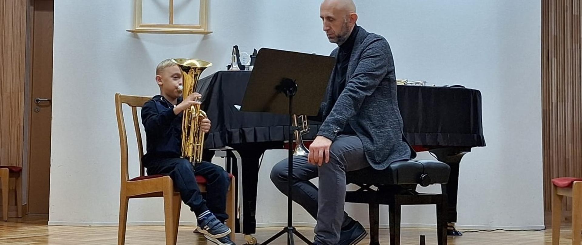 Na scenie w sali muzycznej stoi chłopiec grający na tubie obok dorosłego mężczyzny, również grającego na trąbce. Za nimi znajduje się fortepian, krzesła i pulpit z nutami.
