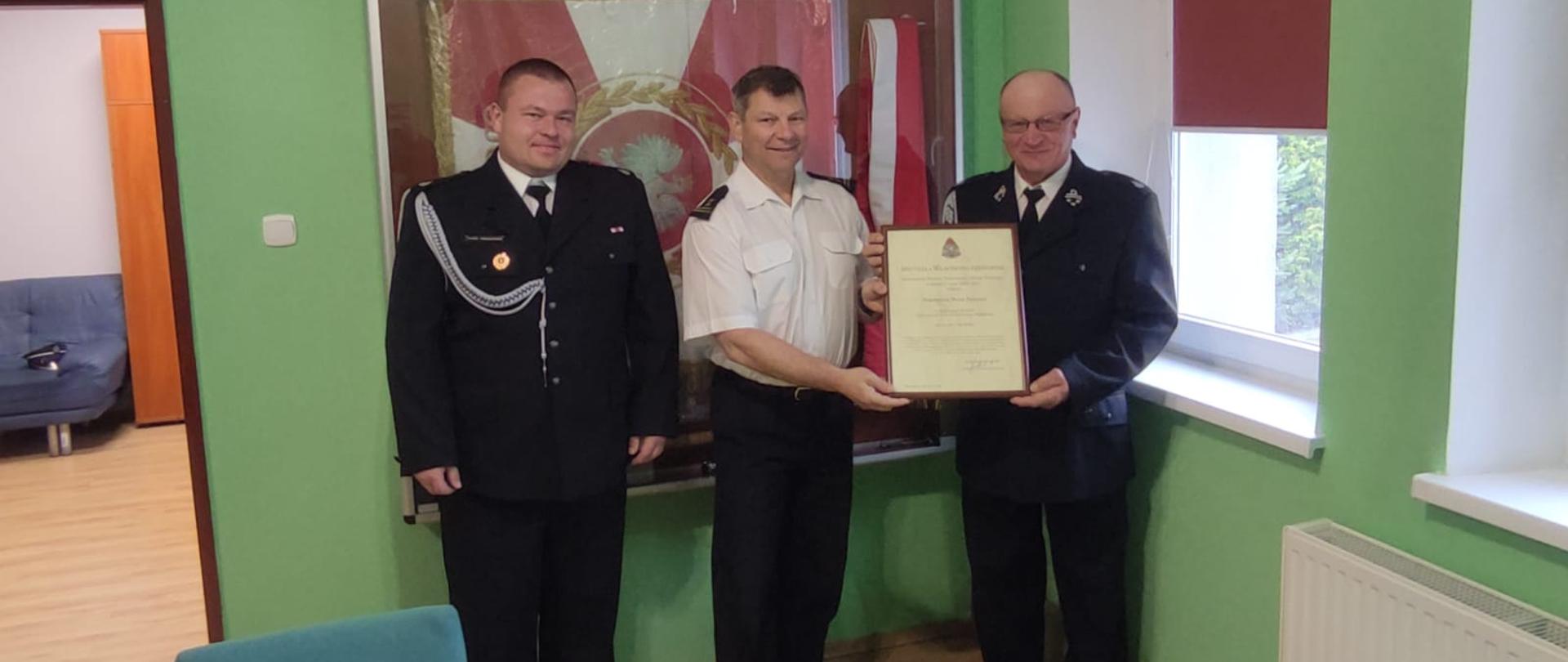 Obraz przedstawia Komendanta Powiatowego oraz 2 strażaków pozujących do wspólnego zdjęcia z przekazaną decyzją. Strażacy w sali odpraw, pora dzienna.