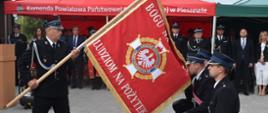 Mężczyzna ubrany w mundur wyjściowy Ochotniczej Straży Pożarnej trzyma sztandar i pochyla go w stronę trzech klęczących mężczyzn ubranych w mundury wyjściowe Ochotniczej Straży Pożarnej. W tle widać zaproszonych na uroczystość gości.