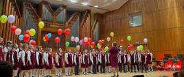 na scenie filharmonii dzieci stojące w półokręgu w czerwonych ubraniach a nad nimi kolorowe baloniki