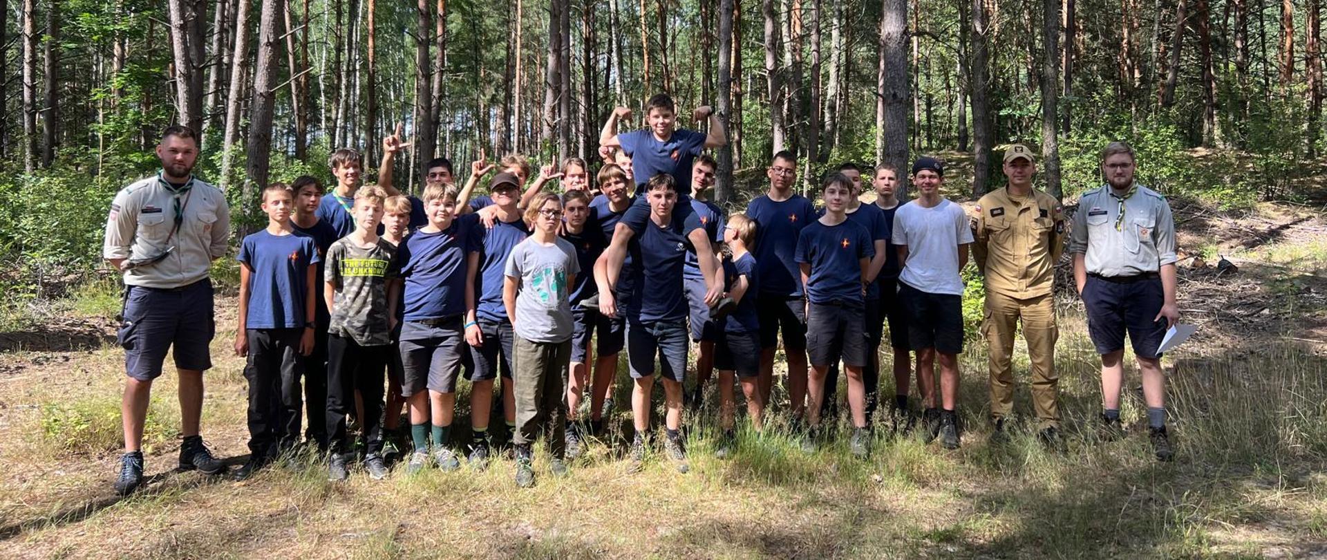 Odwiedziny obozu harcerskiego w ramach akcji „Bezpieczne Wakacje” w miejscowości Rytele Święckie, gm. Kosów Lacki 05.07.2024r. - grupa harcerzy i strażak stoją w dwuszeregu w lesie do zdjęcia