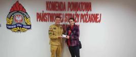 Czujka na straży Twojego bezpieczeństwa. Zdjęcie przedstawia na tle ściany z czerwonym napisem Komenda Powiatowa Państwowej Straży Pożarnej w Rawiczu i logiem PSP, komendanta powiatowego PSP oraz kobietę, która w konkursie wygrała czujnik tlenku węgla. Komendant ma na sobie mundur w kolorze piaskowym. Laureatka ma przewieszoną przez prawe ramię, czarną torebkę. Wręczający i obdarowana trzymają w rękach biały czujnik czadu. 