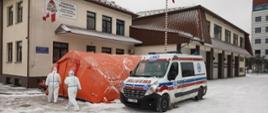 Na zdjęciu widoczny utworzony przez strażaków z Komendy Powiatowej Państwowej Straży Pożarnej w Gorlicach punkt pobrań wymazów do badań nauczycieli w kierunku SARS-CoV-2. Namiot pomarańczowy przed namiotem karetka z Małopolskiej Kolumny Transportu Sanitarnego z Tarnowa i dwaj pracownicy w białych kombinezonach na tle budynku Komendy powiatowej Państwowej Straży Pożarnej w Gorlicach. 