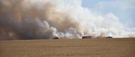 Zdjęcie przedstawia palące się pole, duże zadymienie. Obok stoi wóz strażacki i maszyny rolnicze