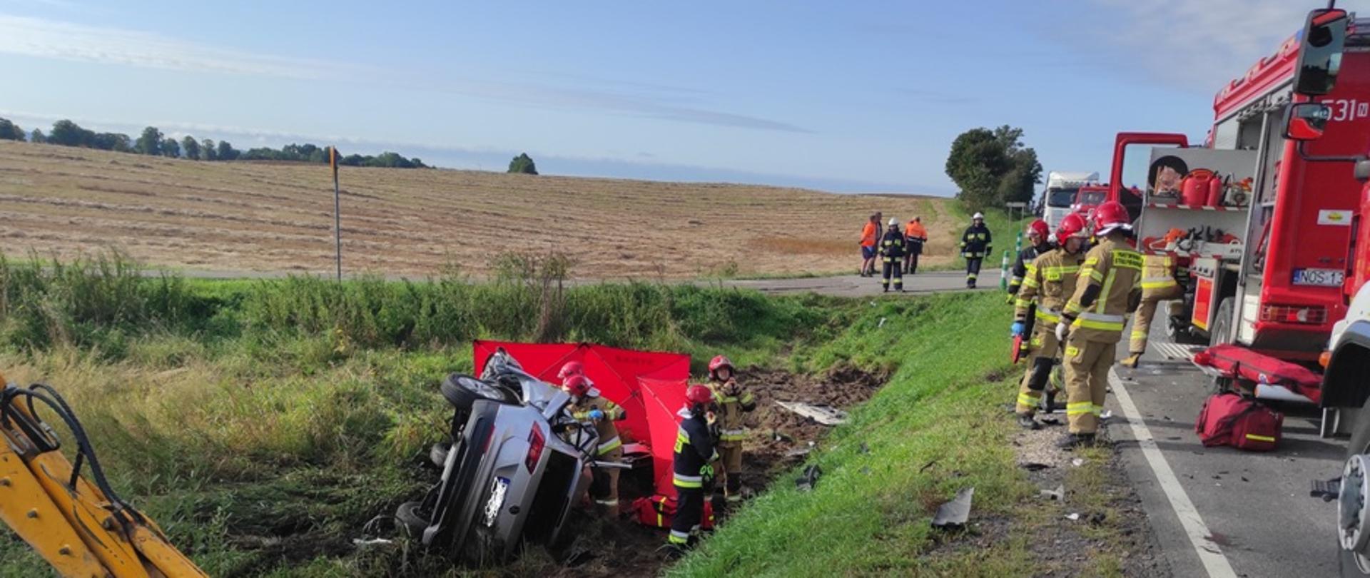 Rozbity samochód osobowy leży w rowie. Strażacy osłaniają parawanem drastyczny widok