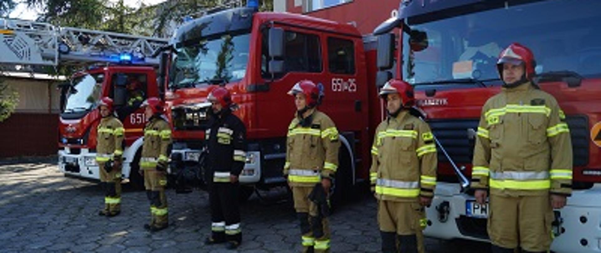 Strażacy w mundurach stojący w odległości 2 metrów od siebie na baczność na tle trzech wozów bojowych