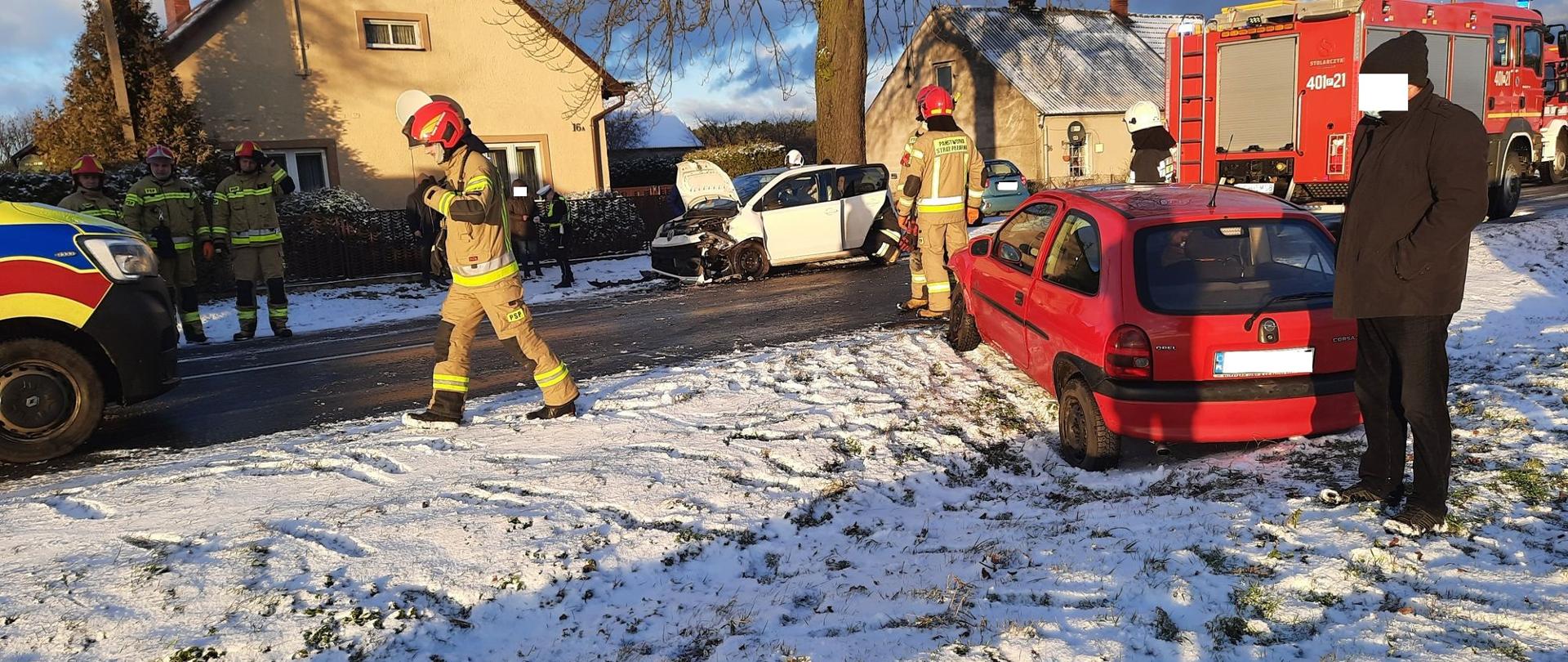 Zdjęcie przedstawia strażaków i policjanta działających przy wypadku drogowym.
W tle pojazdy uszkodzone, ulica i pola.
