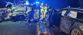 Zdjęcie przedstawia dwa samochody osobowe po wypadku. Jedno auto leży na boku, drugie auto stoi na kołach z uszkodzonym przodem. Na zdjęciu widać samochody strażackie oraz strażaków. Przy jednym z aut leży torba medyczna PSP. Pora nocna. 