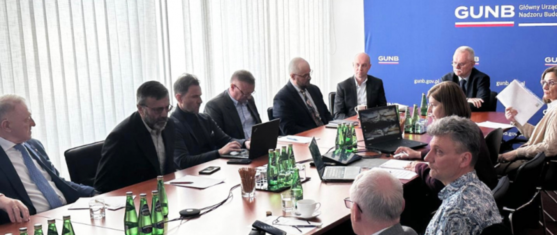 Posiedzenie Rady Wyrobów Budowlanych w Głównym Urzędzie Nadzoru Budowlanego. Uczestnicy spotkania siedzą przy stole, korzystając z laptopów i dokumentów. W tle widoczne jest logo GUNB, a na stole znajdują się butelki wody i inne akcesoria.