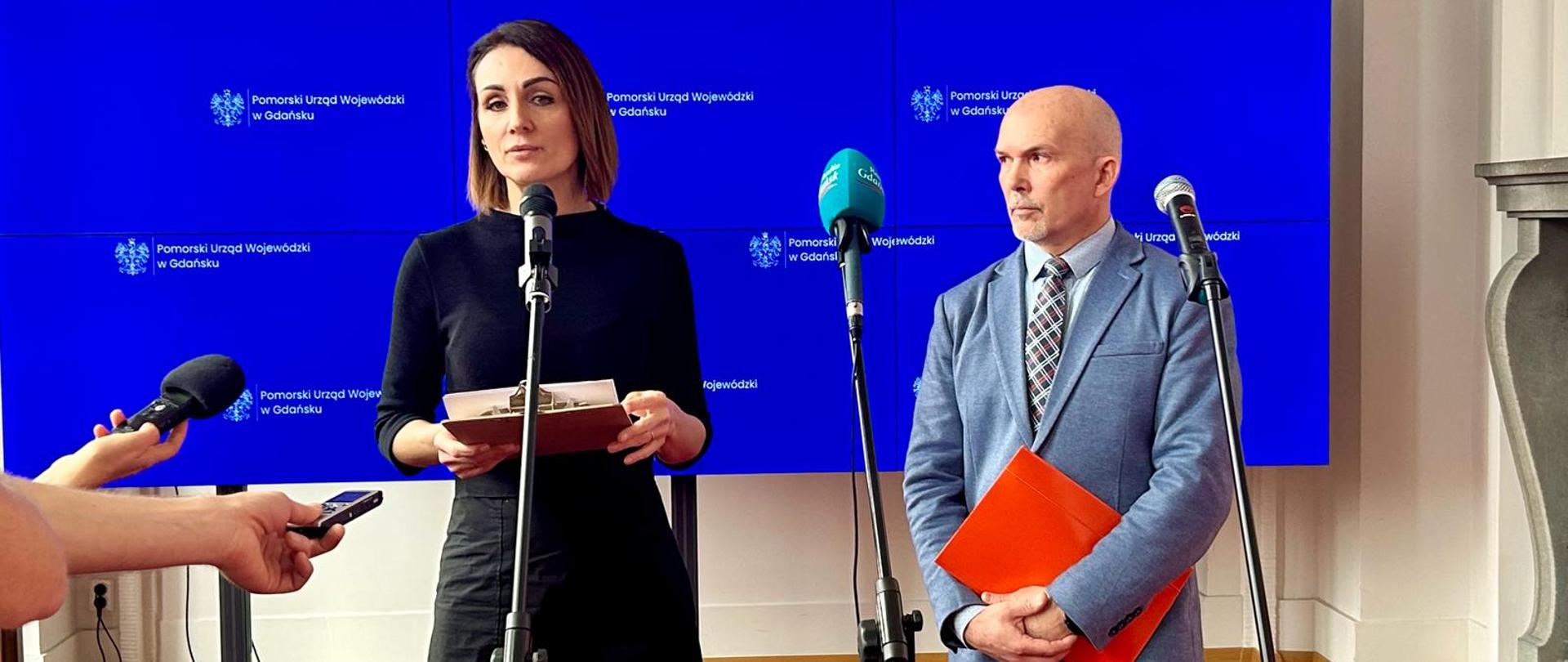 Kobieta (wojewoda) i mężczyzna (konserwator zabytków) stoją przed mikrofonami w dużej sali. Za nimi tablica na które widnieje napis pomorski urząd wojewódzki w Gdańsku