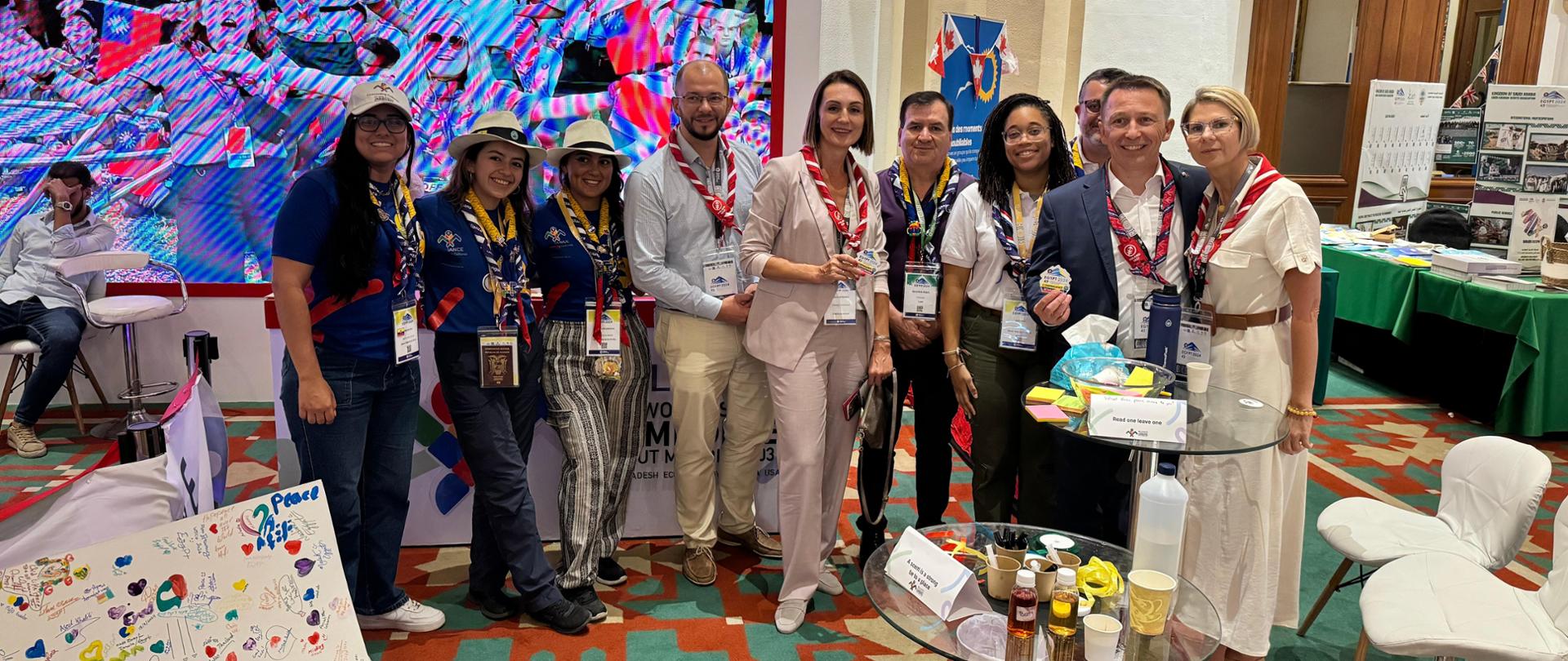 Grupa osób uśmiechających się i stojących razem na tle stoiska podczas 43. Światowej Konferencji Skautowej. Jedną z osób jest wojewoda pomorska.
