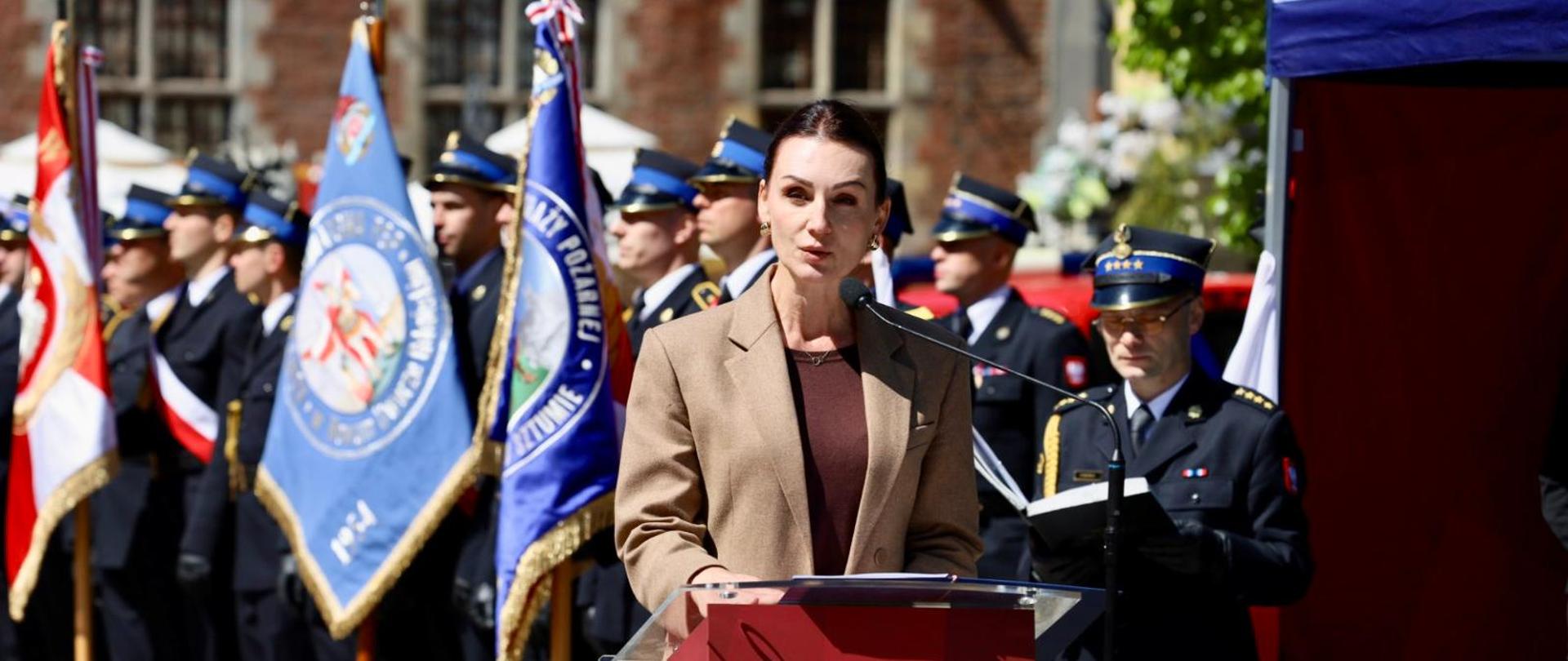 kobieta stoi przy pulpicie i przemawia. W tle stoją strażacy ze sztandarami