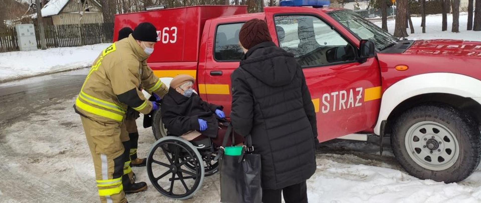 Zdjęcie przedstawia pojazd terenowy należący do jednostki Ochotniczej Straży Pożarnej. Pojazd w kolorze czerwonym z białymi elementami zabudowy. Przednie prawe drzwi pojazdu są otwarte. Przed pojazdem po lewej stronie stoi dwóch strażaków. Obok nich znajduje się wózek inwalidzki, na którym siedzi starsza osoba. Przed nimi stoi jeszcze jedna kobieta, patrząca w stronę strażaków i osoby na wózku. Za pojazdem widać drzewa. Ziemia pokryta jest śniegiem.